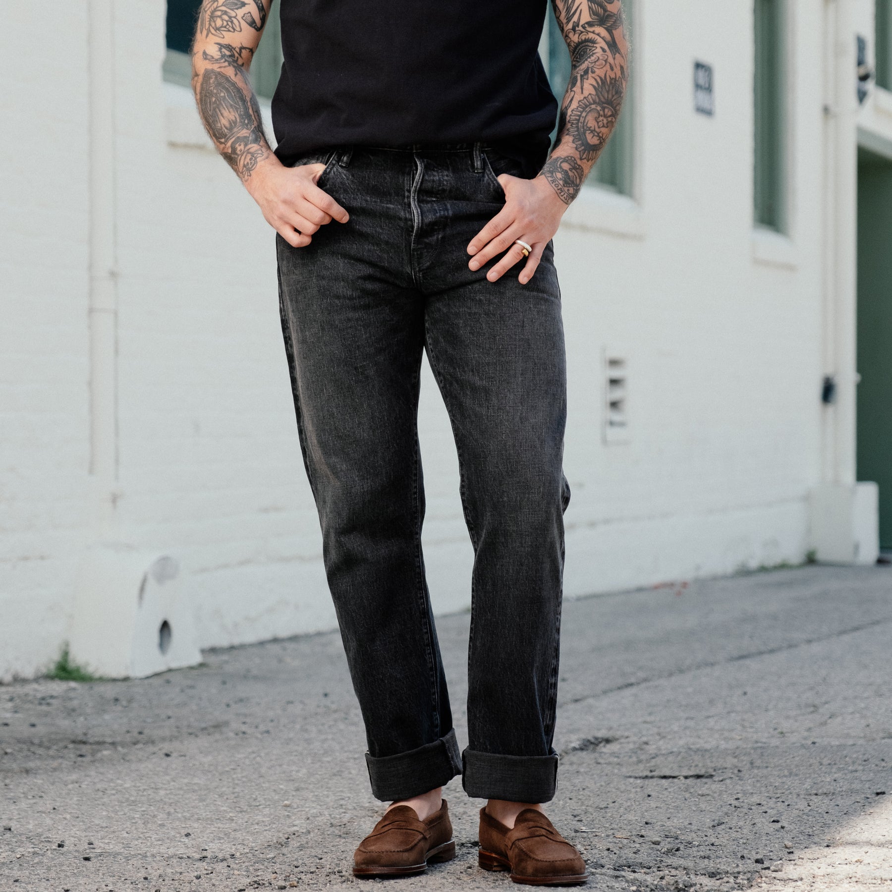 A person wearing dark charcoal gray straight-leg jeans with rolled cuffs, paired with a black t-shirt and brown suede loafers, standing outdoors against a white brick wall.
